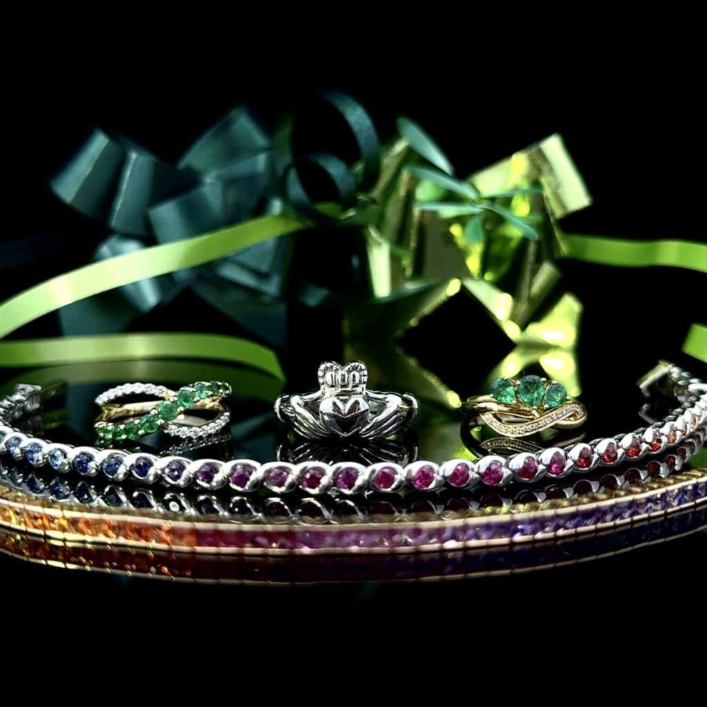 Rainbow Sapphire bracelets stacked in front of 3 rings. A wavy gold ring with green sapphires and diamonds, A white gold Claddagh ring, and a gold ring with large emeralds and a wavy line of accent diamonds