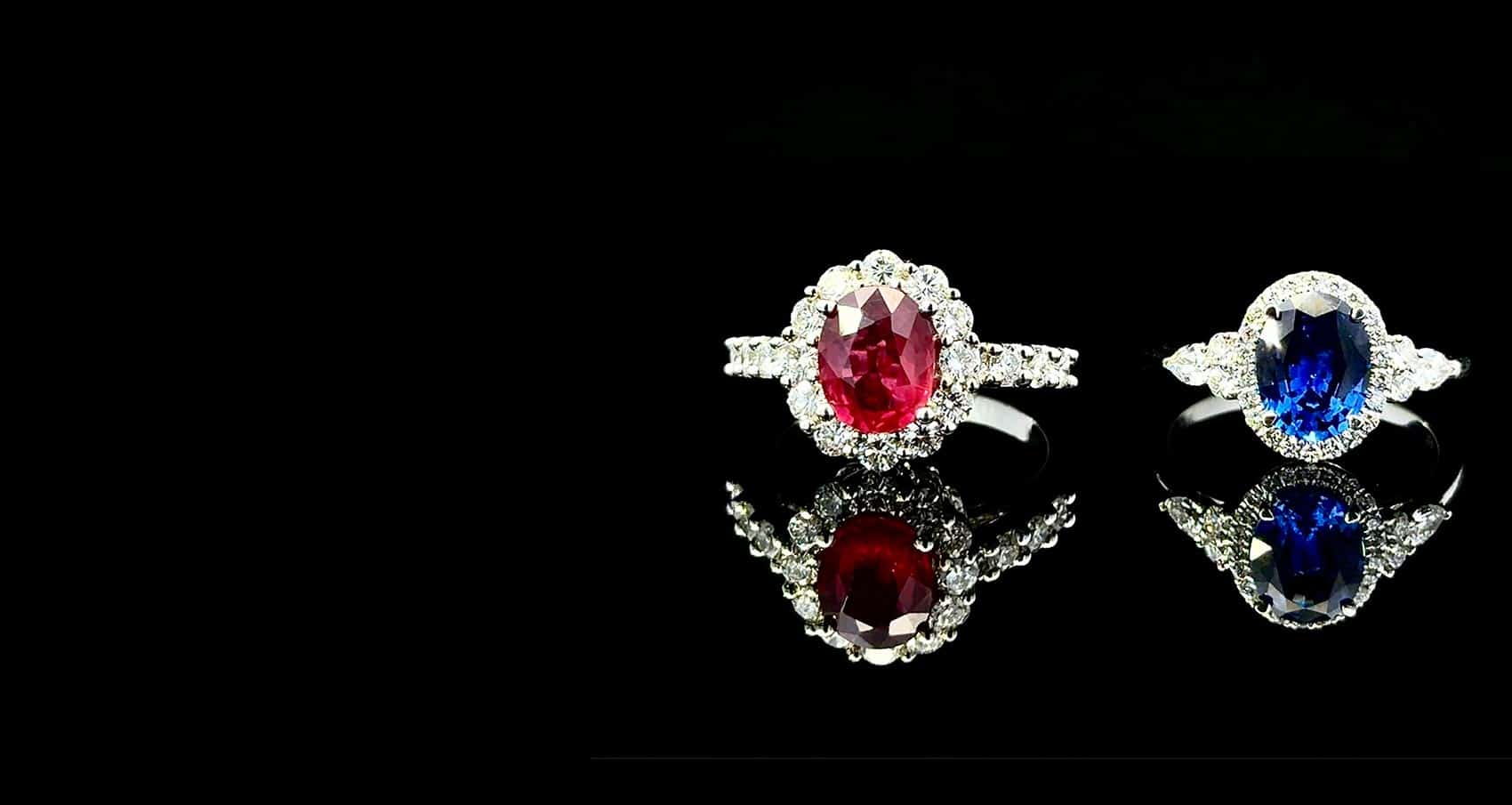 2 rings sitting side by side on a black background. One is a Large red ruby surrounded by diamonds in a white gold ring, the other is a large Sapphire surrounded by diamonds